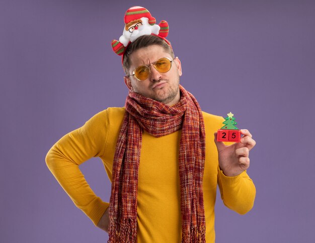 Dissatisfied young man in yellow turtleneck with warm scarf and glasses wearing funny rim with santa on head showing toy cubes with number twenty five standing over purple wall