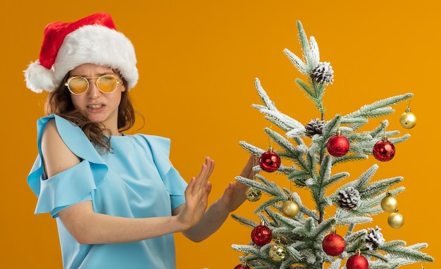 Dissatisfied young woman in blue top and santa hat wearing yellow glasses 