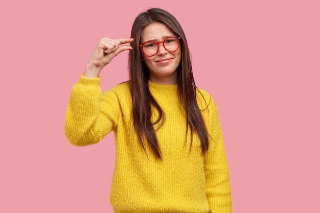 불만족스러운 젊은 여성은 긴 머리를 가지고 있고, 손가락으로 크기를 설명하고, 작은 것을 보여주고, 광학 안경을 쓰고, 노란색 따뜻한 스웨터를 입습니다.