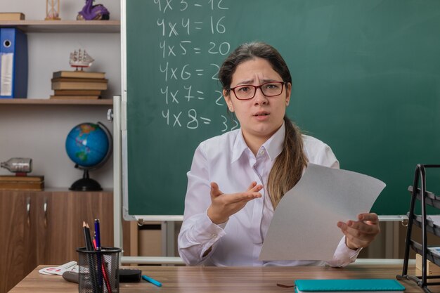 Dissatisfied young woman teacher wearing glasses sitting at school desk with blank pages checking home work looking displeased in front of blackboard in classroom