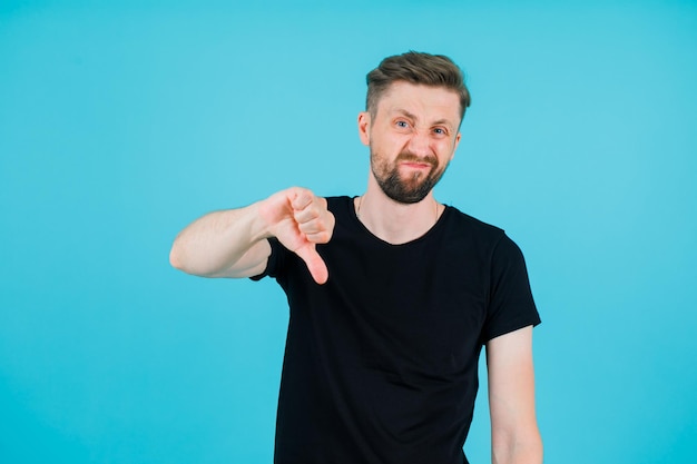 Dissatisfied youngman is showing bad gesture by pointing down with thumb on blue background