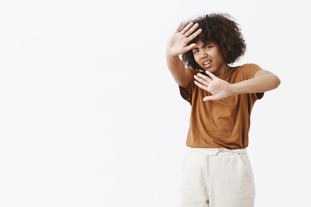 Disspleased annoyed and pissed african american young woman with afro hairstyle protecting from bright light with raised palms covering face and frowning from discomfort