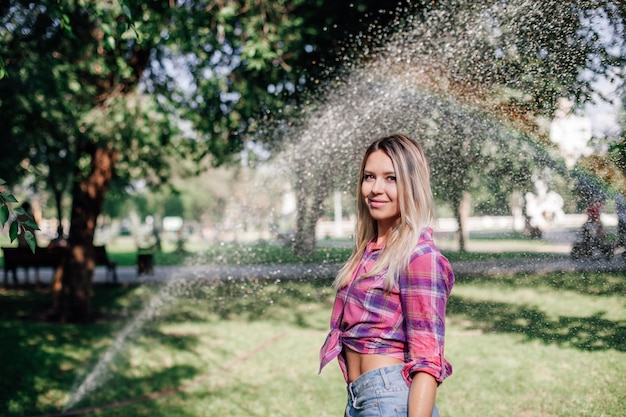 Distant portrait photo of adult shining blonde woman standing in park wearing checkered shirt tied i...