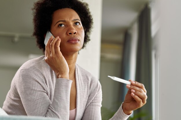 Distraught African American woman communicating over mobile phone while measuring her temperature at home