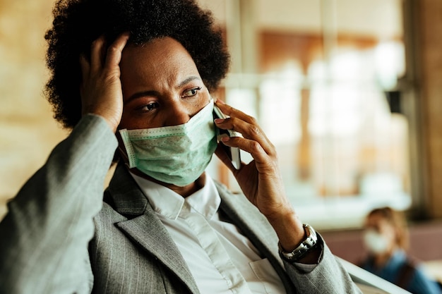 Distraught black businesswoman talking on mobile phone while wearing protective face mask at railroad station hallway