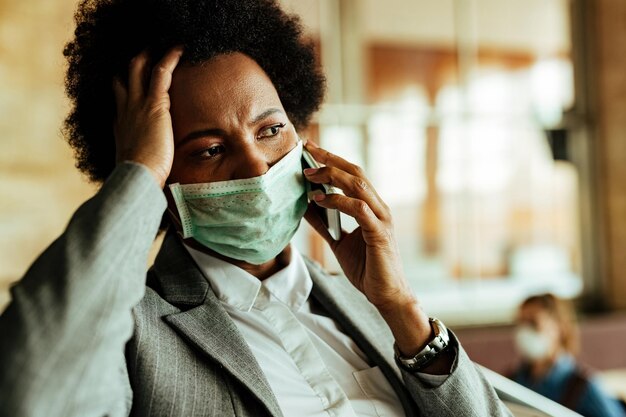 Distraught black businesswoman talking on mobile phone while wearing protective face mask at railroad station hallway