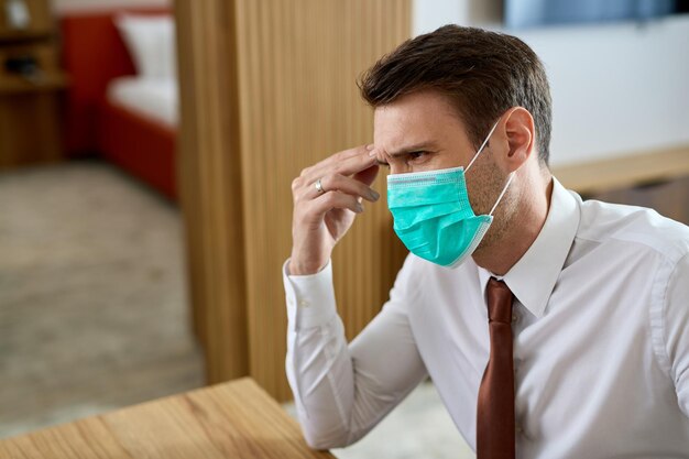 Distraught businessman wearing protective face mask while sitting in hotel room and thinking of something