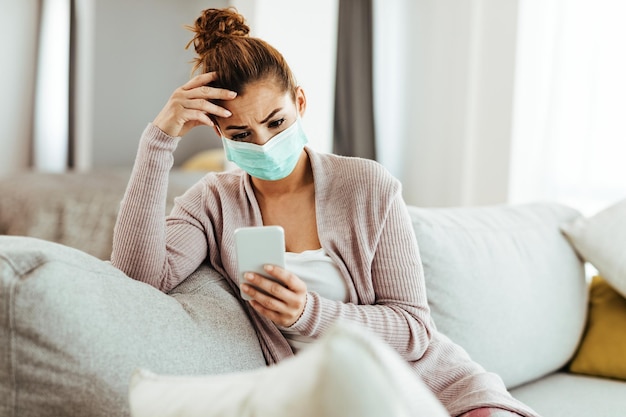 Distraught woman wearing protective mask while sitting on the sofa and text messaging on mobile phone at home