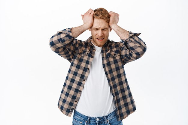 Free photo distressed adult redhead man having headache, holding hands on head and looking upset, disappointed or frustrated of failure, standing over white wall