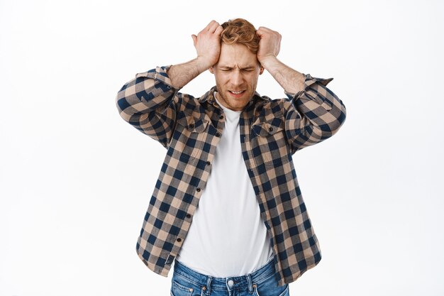 distressed adult redhead man having headache, holding hands on head and looking upset, disappointed or frustrated of failure, standing over white wall