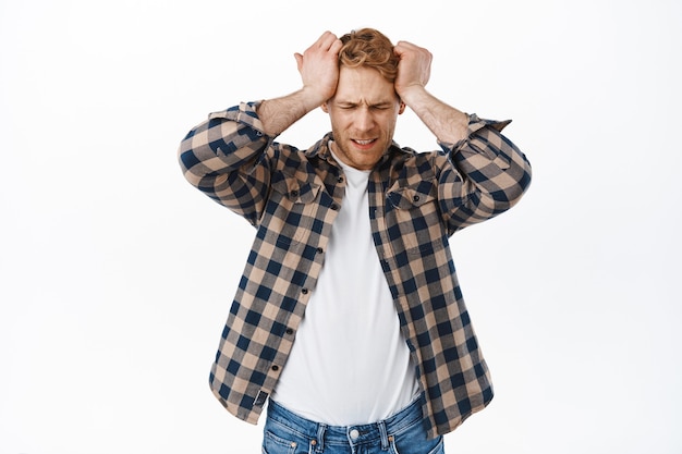 Distressed adult redhead man having headache, holding hands on head and looking upset, disappointed or frustrated of failure, standing over white wall