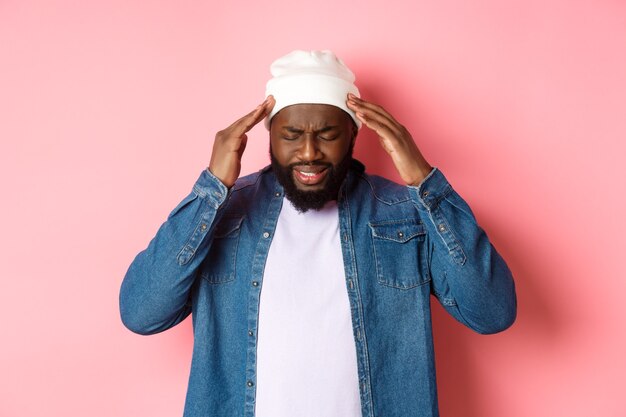 Distressed Black man having headache, touching head and grimacing with concerned face, standing over pink background