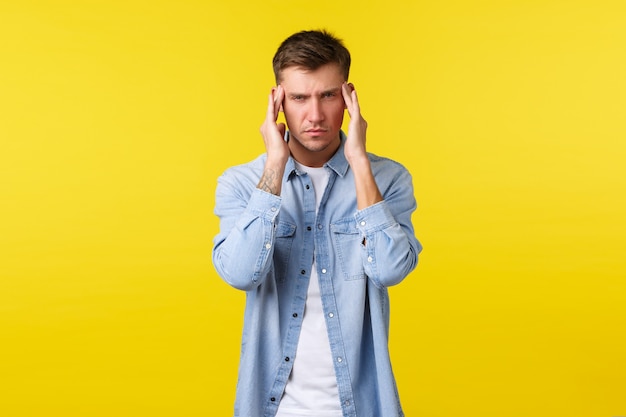 Distressed handsome blond man feeling stress and fatigue. Guy suffering headache, touching heach and grimacing from pain. Man with migraine rubbing temples, trying focus, yellow background.