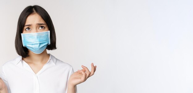 Distressed and miserable young asian woman in face mask looking up looking up sad standing over whit