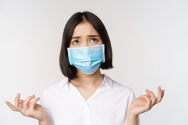 Distressed and miserable young asian woman in face mask looking up looking up sad standing over white background