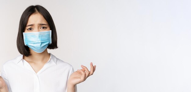 Distressed and miserable young asian woman in face mask looking up looking up sad standing over white background