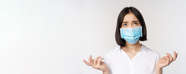 Distressed and miserable young asian woman in face mask looking up looking up sad standing over white background