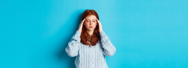 Distressed teenage redhead girl touching head looking down with troubled face expression standing ag