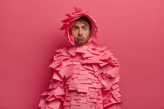 Free photo distressed unhappy man purses lower lip, looks with offended expression, wears outfit made of adhesive notes, poses against pink wall, dissatisfied with something, being in low spirit.