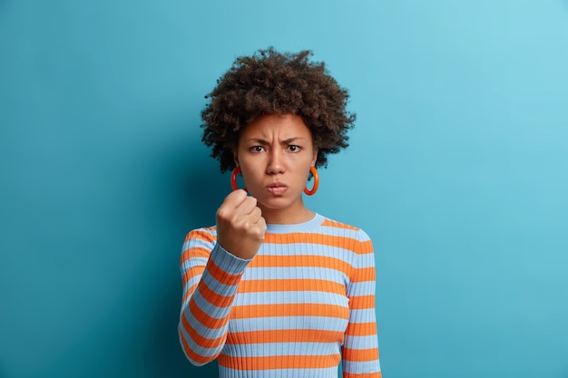 La donna arrabbiata disturbata stringe il pugno e ha un'espressione indignata, avverte di vendetta, esprime odio ed emozioni negative, vuole un pugno al nemico, vestita in modo casual, isolata sul muro blu.