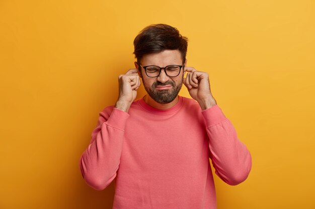 Disturbed bothered man plugs fingers at ear holes, hears annoying loud sound, noise or siren, frowns face, dressed casually, poses over yellow wall, ignores heartbreaking scream coming from other room