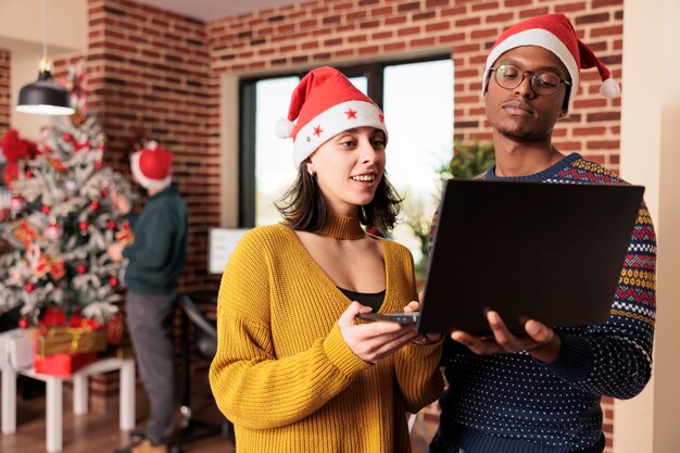 冬のホリデーシーズンにクリスマスの飾りやライトで飾られた空間で働いて、お祝いのオフィスでコンピューターを見ている多様な同僚。クリスマスイブに仕事でサンタの帽子をかぶっている人。