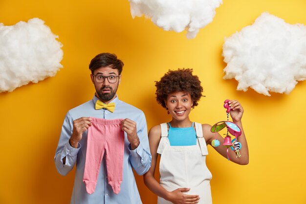 Diverse couple expect baby pose with toys and clothes for child going to become parents.