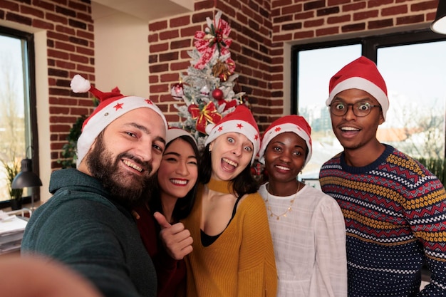 Разнообразные коллеги фотографируются на праздничном рабочем месте, празднуют рождественский праздник с елкой и украшениями. Люди фотографируются с сезонными украшениями в бизнес-пространстве.