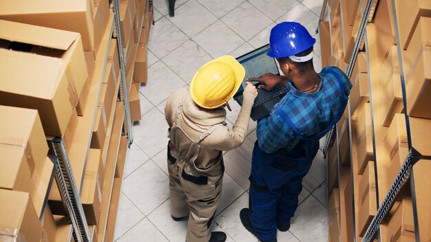 Diverse employees analyzing inventory list on laptop, counting products in boxes on storage room shelves. Young people using device to work on stock logistics, delivery service.