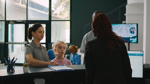 Free photo diverse family coming to facility reception desk in lobby, talking to receptionist about checkup appointment with medic. little girl and parents waiting to attend consultation, waiting area.