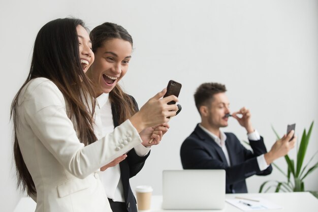 Diverse female colleagues laughing having fun with smartphone in office