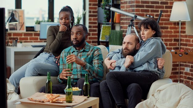 Diverse friends being scared watching horror movie, doing jump scare and feeling terrified of scary thriller film on tv channel program. Getting frightened and afraid, closing eyes in shock.