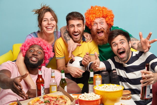 Diverse friends football supporters celebrate success of favorite team with popcorn, pizza and drinks, sit on couch, spend Sunday evening in front of TV, isolated over blue wall. Home cinema