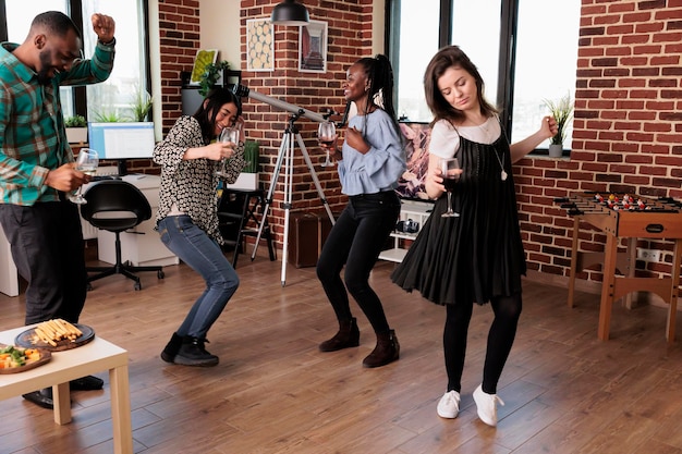 Diverse group of close friends dancing in living room while celebrating birthday event. Multicultural people relaxing with music while enjoying wine party at home together.