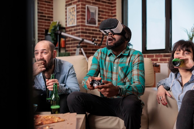 Diverse group of friends using virtual reality headset to play video games on television. Having fun with beer bottles and snacks at house party, playing competition with vr goggles.