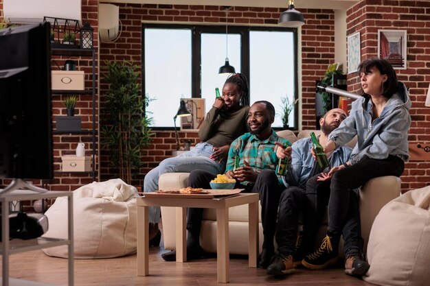 Diverse group of friends watching movie on television, eating popcorn and drinking bottles of beer at fun gathering. People looking at film on tv channel program, enjoying leisure activity.