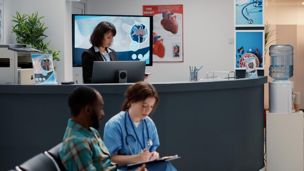 Diverse group of patients asking for help at hospital reception desk, sitting in waiting area lobby. Mother with child, senior man and asian patient having appointment at busy medical clinic.