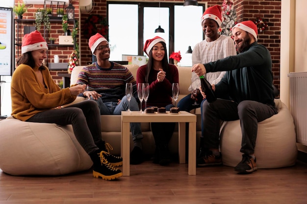 オフィスパーティーでクリスマスを祝い、グラスでワインを飲み、冬のホリデーシーズンを楽しんでいる人々の多様なグループ。クリスマスツリーと季節の装飾、アルコール飲料でお祭りの職場。