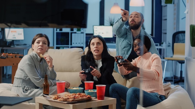 Разнообразная группа людей, играющих в видеоигры на телевизионной консоли после работы. Коллеги наслаждаются игрой с контроллерами по телевизору, чтобы развлечься и развлечься с помощью технологий.