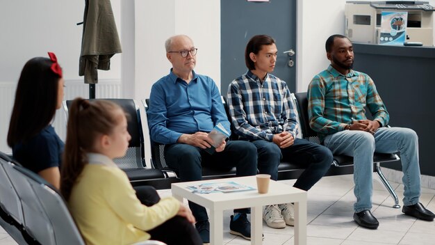 Diverse group of people sitting together in hospital waiting room, preparing to meet with general practitioner at medical clinic. Different patients waiting to attend appointment. Tripod shot.