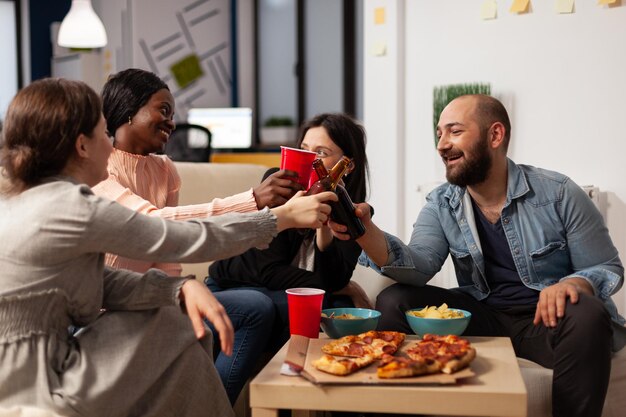 Diverse group of workmates clinking glasses and bottles at office celebration with drinks and snacks in office. Colleagues celebrating with alcoholic beverage, making cheers toast.