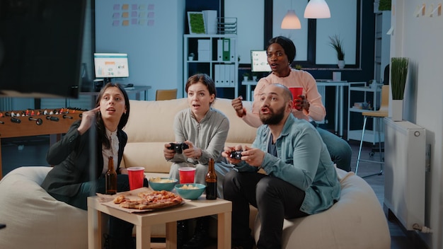 Diverse group of workmates using joysticks to play video games on television. Colleagues cheering while playing game with controllers on tv console to have fun and entertainment.