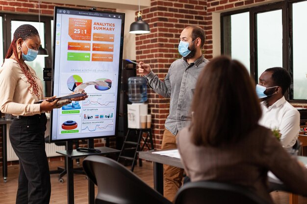 Diverse managers pointing at marketing presentation explaining financial strategy working in startup company office. Businessteam with medical face mask against covid19 brainstorming project ideas