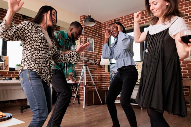 友好記念日を祝いながら、居間で踊る多様な人々。自宅のリビングルームで開催されたワインパーティーで踊りながら、笑顔で一緒に楽しんでいる多民族の友人グループ。