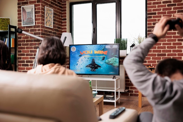 Разные люди сидят в гостиной и наслаждаются играми на электронном развлекательном устройстве. Игривая многонациональная группа друзей сидит на диване и играет в консольные видеоигры с контроллером.