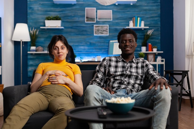 リビングルームのソファで赤ちゃんの子供を期待しながら、家族の映画を見ている多様なリラックスした未来の両親。注意を払いながらアフリカ系アメリカ人男性の会社を楽しんでいる陽気な妊娠中の白人女性