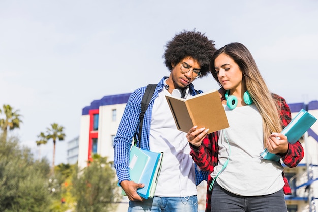 Diverse student couple standing in front of university building reading the book