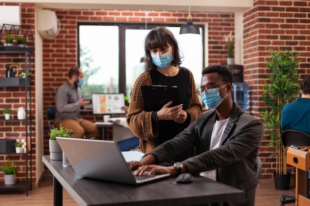 Diverse team analyzing company statistics working at marketing project in brick wall startup office. Multiethnic businesspeople wearing medical face mask to prevent infection with coronavirus