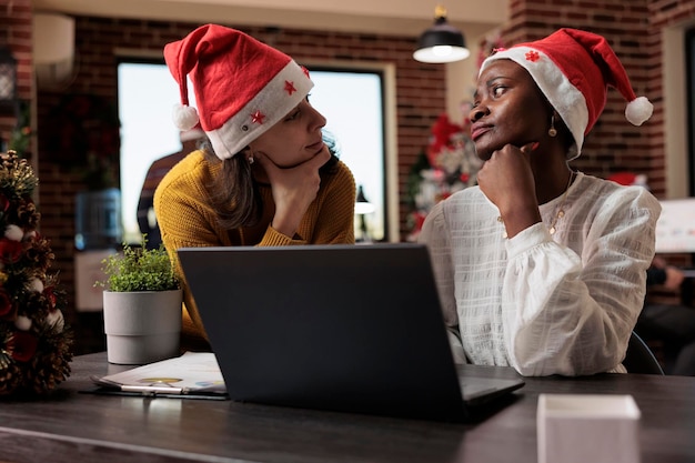 Diverse team of colleagues with santa hat working on business at laptop, sitting in festive office filled with christmas decorations and ornaments. Doing teamwork and celebrating holiday.