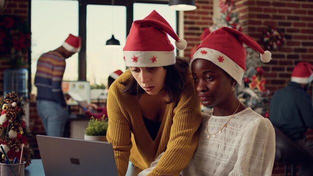 크리스마스 트리와 장식으로 겨울 시즌을 축하하며 축제 사무실에서 보고서 작업을 하는 다양한 동료 팀. 크리스마스 시간에 노트북을 보고 팀워크를 하는 여성.